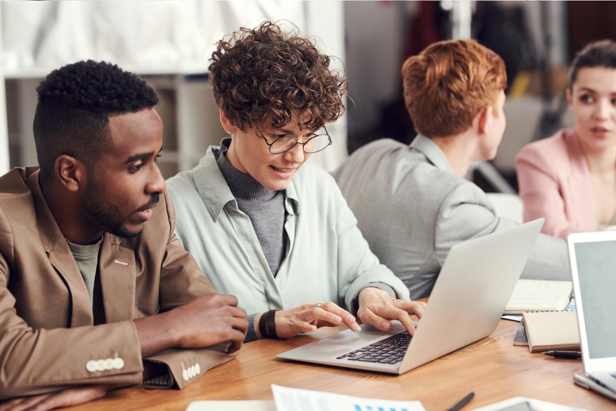 Four people sit at a table, with two focused on a laptop in the foreground. One person is typing while the other watches. The background shows two others engaged in conversation.