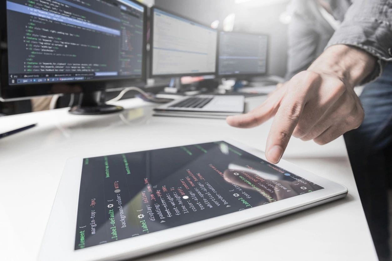 A person interacts with code displayed on a tablet, with multiple computer monitors showing more coding content in the background.