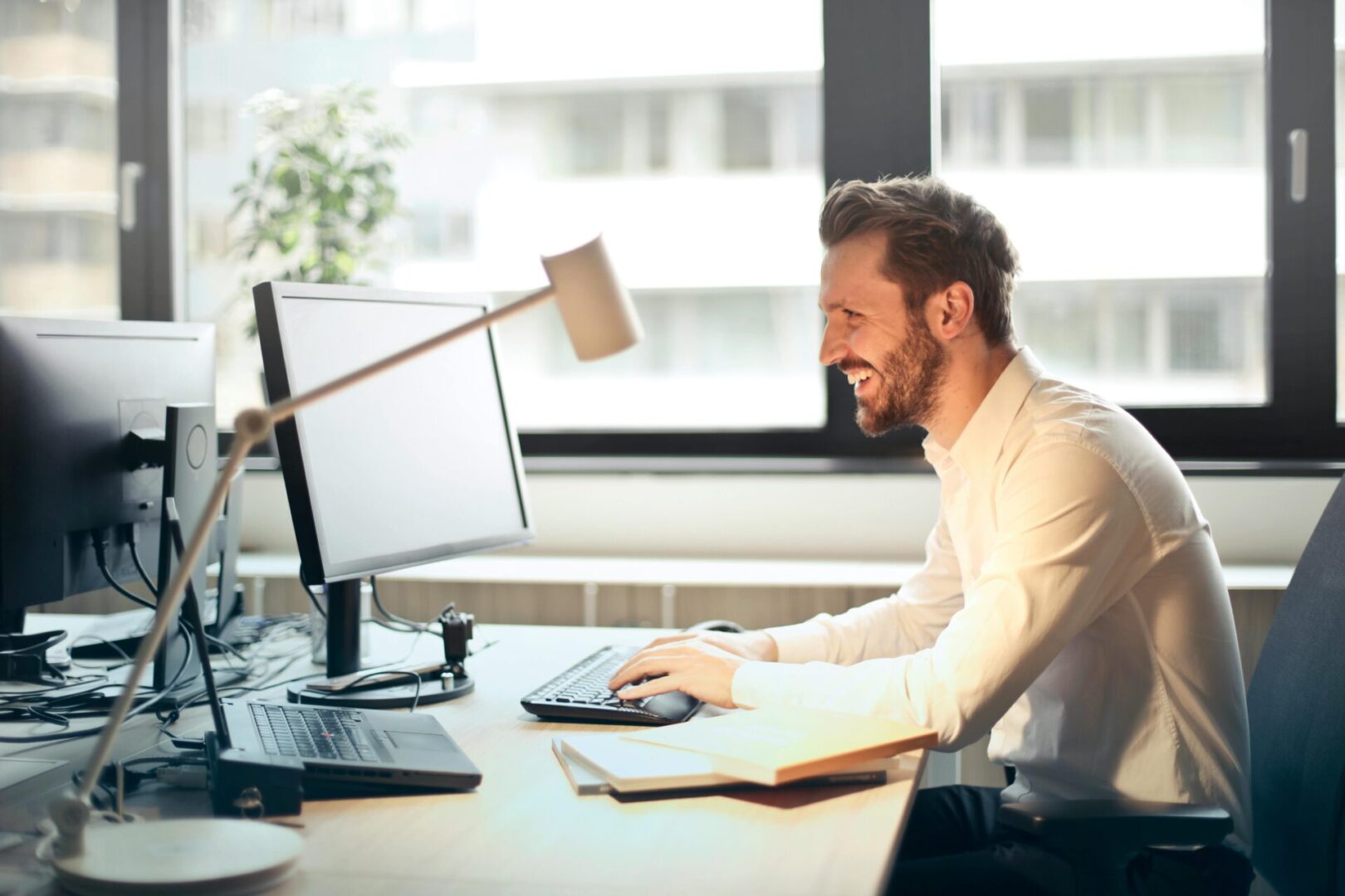 A man in a white shirt sits at a desk working on a computer. He is smiling and there is a desk lamp and some papers beside him. Large windows and a building view are in the background.