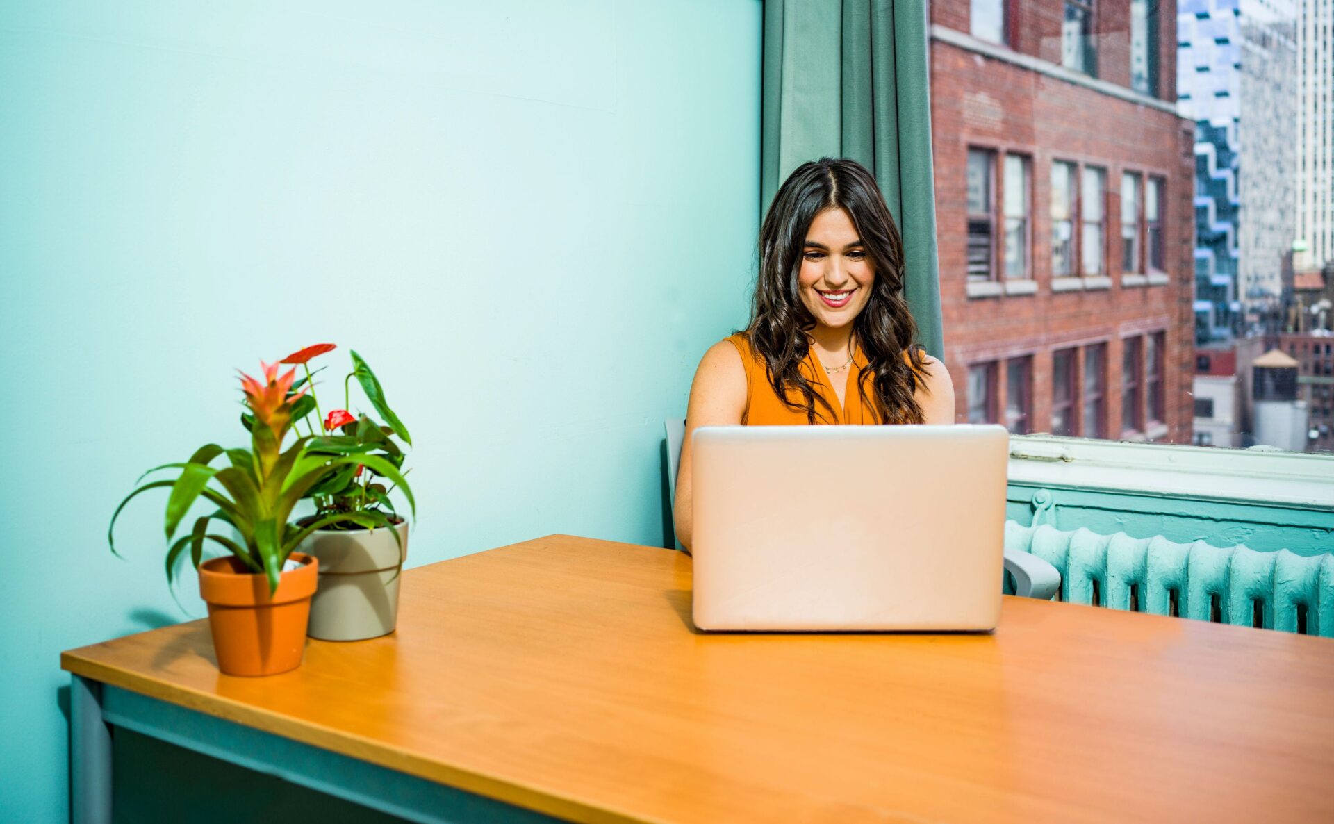 A woman sits at a wooden desk working on a laptop in an office, with two potted plants on her left and a view of buildings through the window behind her.