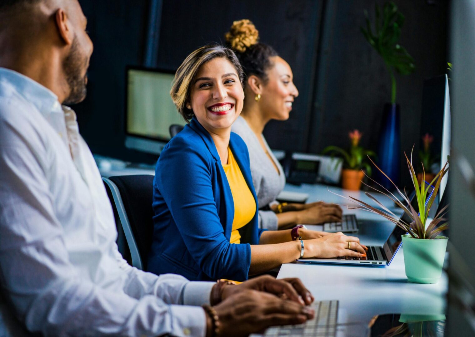 Three people sit at a desk working on laptops, with the woman in the middle smiling and looking towards the person on the left.